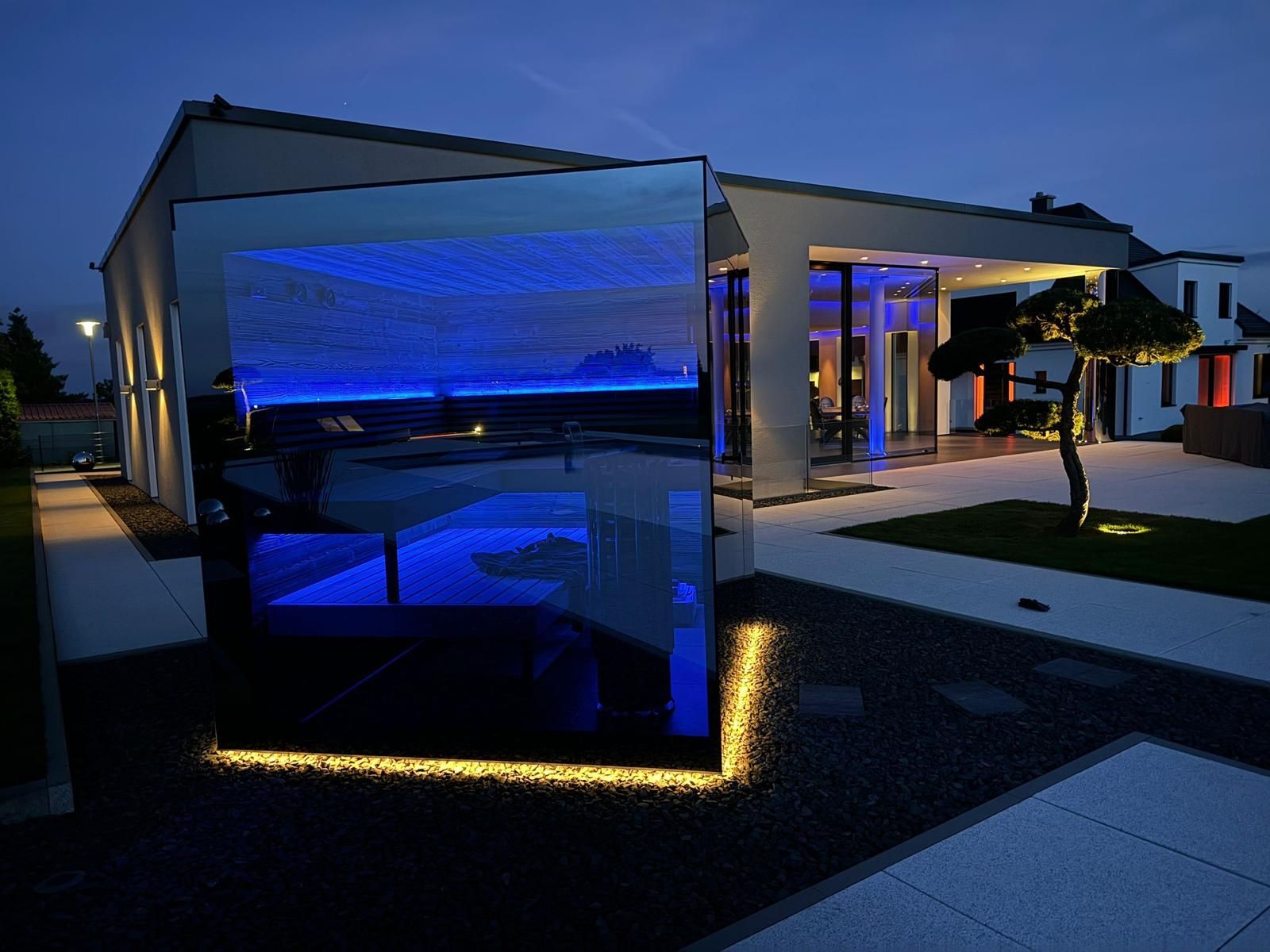 Sauna de vidro com iluminação LED azul, situada num jardim moderno, criando um ambiente sofisticado e relaxante ao anoitecer.Sauna de vidro com iluminação LED azul, situada num jardim moderno, criando um ambiente sofisticado e relaxante ao anoitecer.